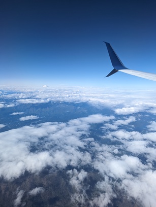 Photo of the window view of an airplane. A wing of the plane can be seen on the side of the image. Below are scattered clouds, and a dark green landscape. In the horizon is a tall, white mountain that breaks through the clouds.