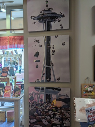 Photo taken inside a bookstore, of art. The picture is split into 3 separate vertical frames. It is of the Space Needle, with pandas crowding below it, on top of it, and even falling from it. Each panda is doing, holding, or wearing something unique that references the city's culture.