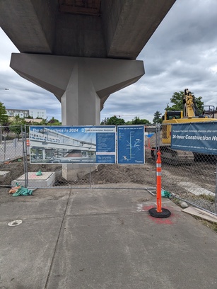 Photo underneath a Light Rail overpass, with construction being done below. On a chain link fence is a sign informing people about the planned rail path. "The train arrives in 2024", it says.