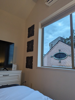 Photo taken inside a bedroom, with a TV on a dresser placed against the wall. The adjacent wall has a large window. Outside is the roof of a neighboring house, and the Space Needle in the distance.