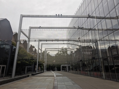 Photo of a long concrete pathway, with strange rope, line-looking things above it. To the right is a tall building almost entirely made of glass windows.