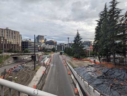 Photo taken from a highway overpass. The parts next to the road are still under construction. In the distance are buildings and evergreen trees.