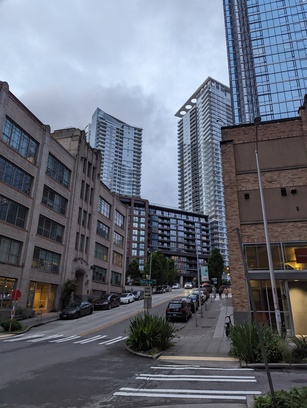 Photo of some buildings a few stories high along a road sloping upwards. Tall, glassy skyscrapers loom in the background, behind the buildings.