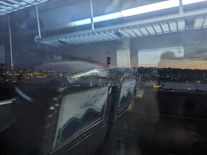 A photo looking out from the bus window. The reflection of bus seats and lights are overlayed on a beautiful view of the Seattle skyline at sunset. You can even see the Space Needle!