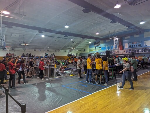 Photo from inside a high school gym turned into a robotics competition, with people from several teams wearing matching shirts crowded in their own spots, with a small fenced-off zone for the robots to fight in. People watch and cheer from the bleachers.