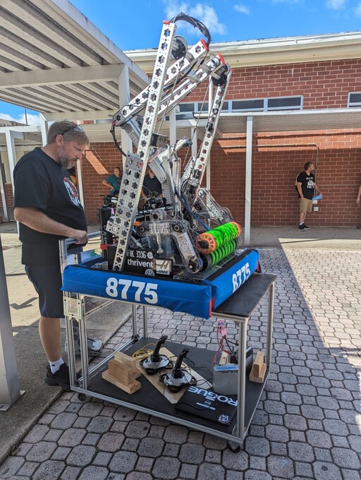 Outdoor photo of my team's robot. Wheeled in on a small cart, a robot labeled 8775, with a complicated design of metal, wires, and various other robot-y components.